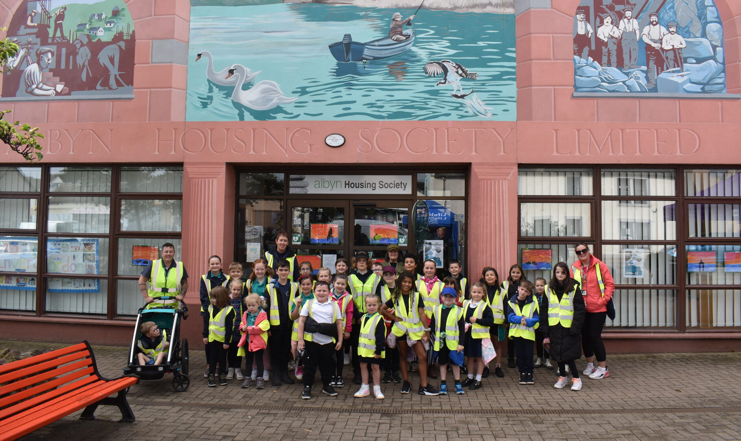 A group of school children stand under a mural of a loch with a person fishing in a boat.