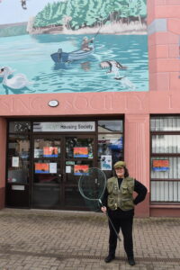 A woman dressed as an angler and holding a fishing net stands under a mural depicting Loch Kildary.