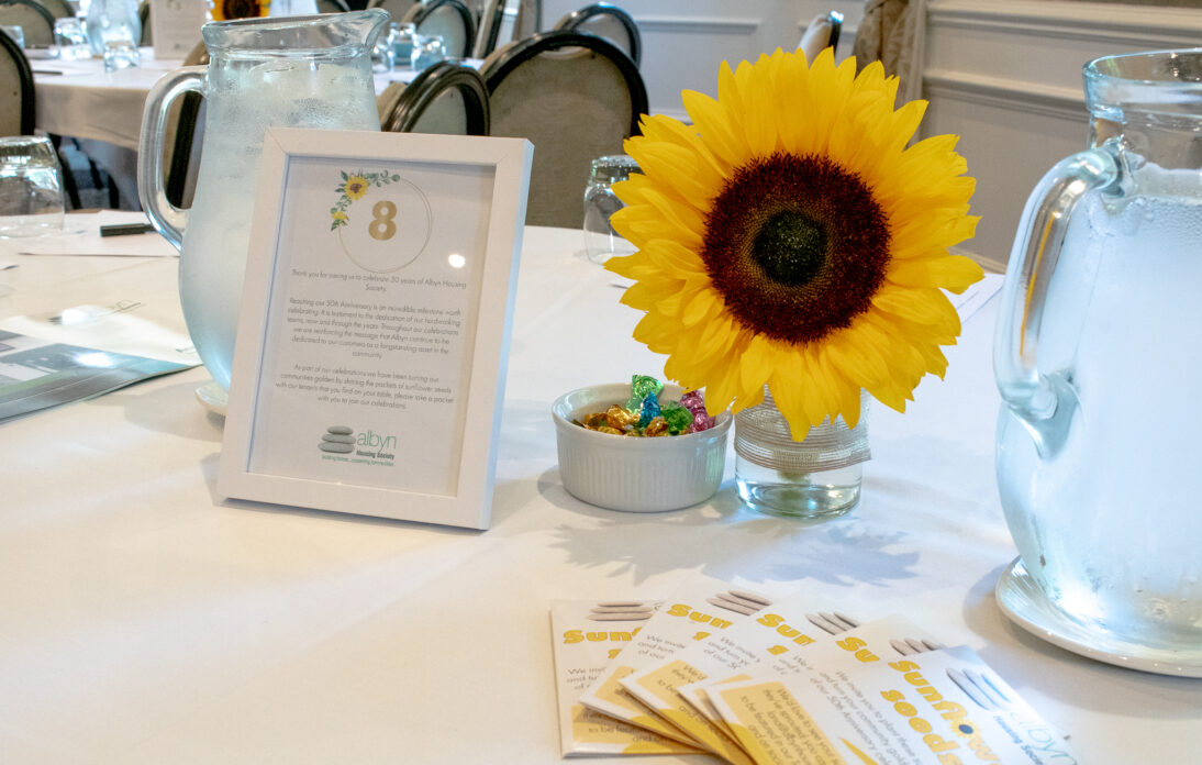 A table is laid out with a photo frame including the table number, a sunflower in a glass vase and some branded sunflower seed packets are on the table.
